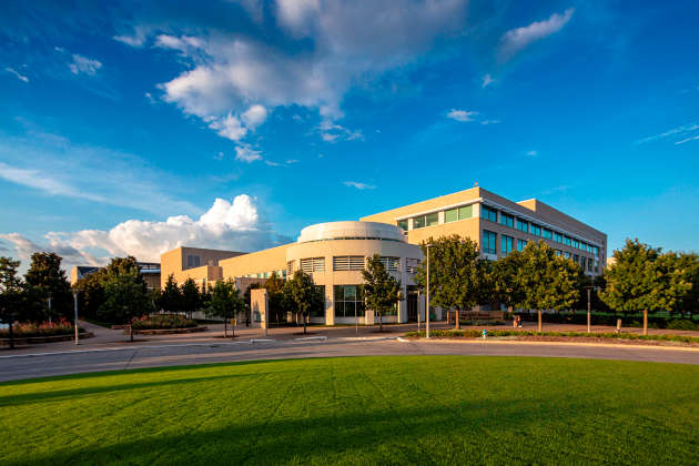 The Naveen Jindal School of Management at UT Dallas sits under a blue sky with green grass leading up to the campus doors.